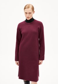 Abito lavorato a maglia bordeaux con maniche lunghe, scollo rotondo e spacchi laterali. Tessuto morbido e vestibilità comoda, con un dolcevita nero sotto.