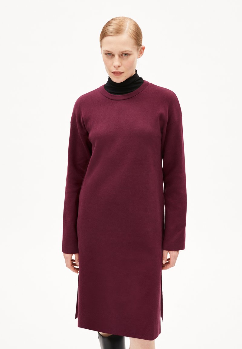 Abito lavorato a maglia bordeaux con maniche lunghe, scollo rotondo e spacchi laterali. Tessuto morbido e vestibilità comoda, con un dolcevita nero sotto.