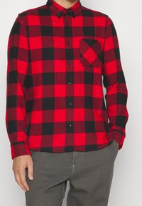 Chemise à manches longues à carreaux rouges et noirs avec un col à boutons, dotée d'une poche poitrine unique et de boutons noirs. Portée avec un pantalon gris.