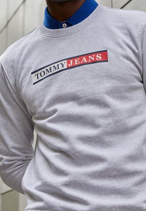 Šedá mikina s dlouhými rukávy, na přední straně s logem "TOMMY JEANS" v červené a modré barvě, kulatý výstřih a bavlněný materiál.