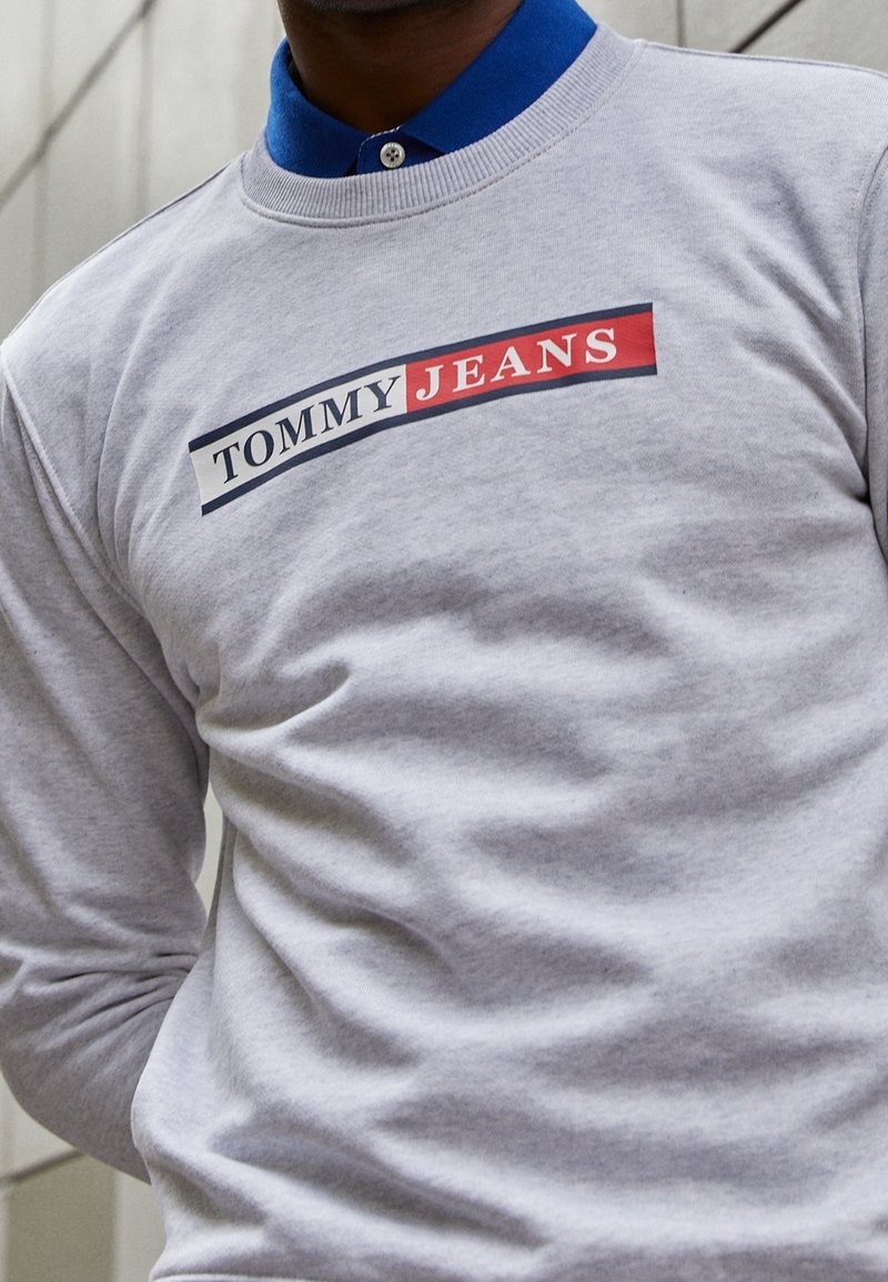 Šedá mikina s dlouhými rukávy, na přední straně s logem "TOMMY JEANS" v červené a modré barvě, kulatý výstřih a bavlněný materiál.
