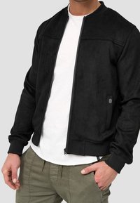 Veste bomber en daim noir avec une fermeture éclair sur le devant, des poignets côtelés et un petit logo sur la poche. Portée sur une chemise blanche.