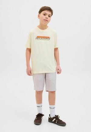 Šviesiai geltona marškinėliai su "JACK & JONES" logotipu, derinami su šviesiai pilkomis šortais. Rudi sportbačiai ir balti šaliko kojinės su juodu užrašu puikiai užbaigia įvaizdį.