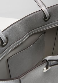 Srebrna torebka wykonana z teksturowanego materiału, z przestronnym wnętrzem i płaską kieszenią. Posiada dwie rączki oraz kontrastowe detale szycia.