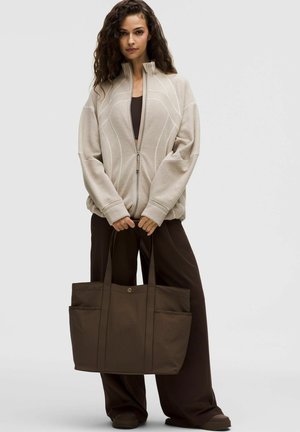 Žena s kovrčavom kosom nosi bež jaknu na patentni zatvarač, tamno smeđe široke hlače i drži veliku tamno smeđu torbu za nošenje.