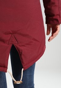 Dobladillo de chaqueta roja texturizada con abertura lateral, cordón ajustable con tope de madera, llevada sobre vaqueros. Primer plano de los detalles.