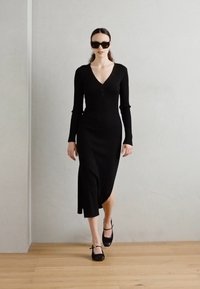 Black long-sleeve knit dress with a deep V-neck and side slit. Worn with black shoes featuring bows. Model wearing black sunglasses, minimalist background.