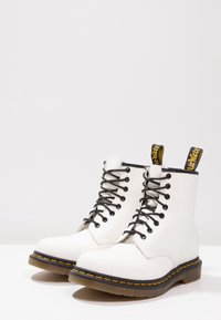 Botas de tobillo de cuero blanco con cordones negros, costuras amarillas y suela de goma negra. Presenta tiradores amarillos para facilitar su uso.