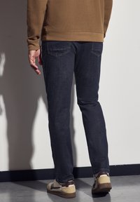 Jeans de mezclilla azul oscuro con corte ajustado, con bolsillos traseros y combinados con zapatillas beige y marrón, de pie frente a una pared clara.