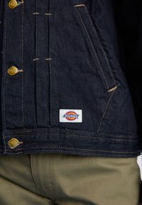 Giacca in denim blu scuro con hardware dorato, chiusure a bottoni e etichetta cucita. Presenta cuciture a contrasto e tasche laterali.