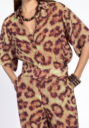 Frau trägt ein braun-beiges, abstrakt gemustertes Hemd mit Knopfleiste und passende Hosen, dazu ein massiver dunkler Halsreif und Armbänder.