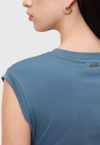 T-shirt blu ribbed con maniche corte, visto da dietro. Presenta un piccolo etichetta al collo e un orecchio di una persona adornato con orecchini esagonali.