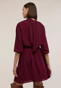 Bordeaux Kleid mit rundem Ausschnitt und gerafftem Stoff auf der Rückseite, ausgestattet mit Ballonärmeln und einem gegurteten Taillieren; glatte, fließende Textur.