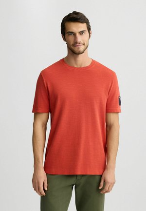 Rotes Kurzarm-T-Shirt mit Rundhalsausschnitt und subtiler Struktur, kombiniert mit grünen Hosen. Akzentuiert durch ein Logo auf dem Ärmel.