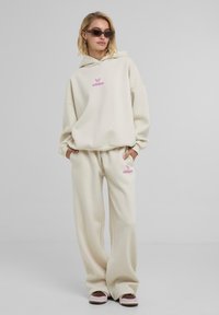 Kreemivärvi oversize-kapütsiga spordijakk ja sobivad dressipüksid, millel on roosa logo tikand. Pehme, fleece-sarnane materjal ja mugav lõige.