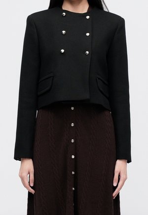 Zwarte cropped jas met zilveren knopen en klepzakken, gedragen over een donkerbruine rok met knopen en een gestructureerd breipatroon.