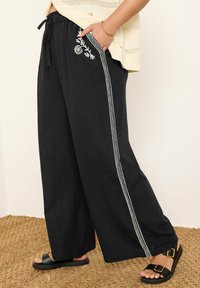 Svarta vida byxor i mjukt tyg med vit brodyr och sidostrick. En dragsko i midjan och sidfickor ger funktionalitet.