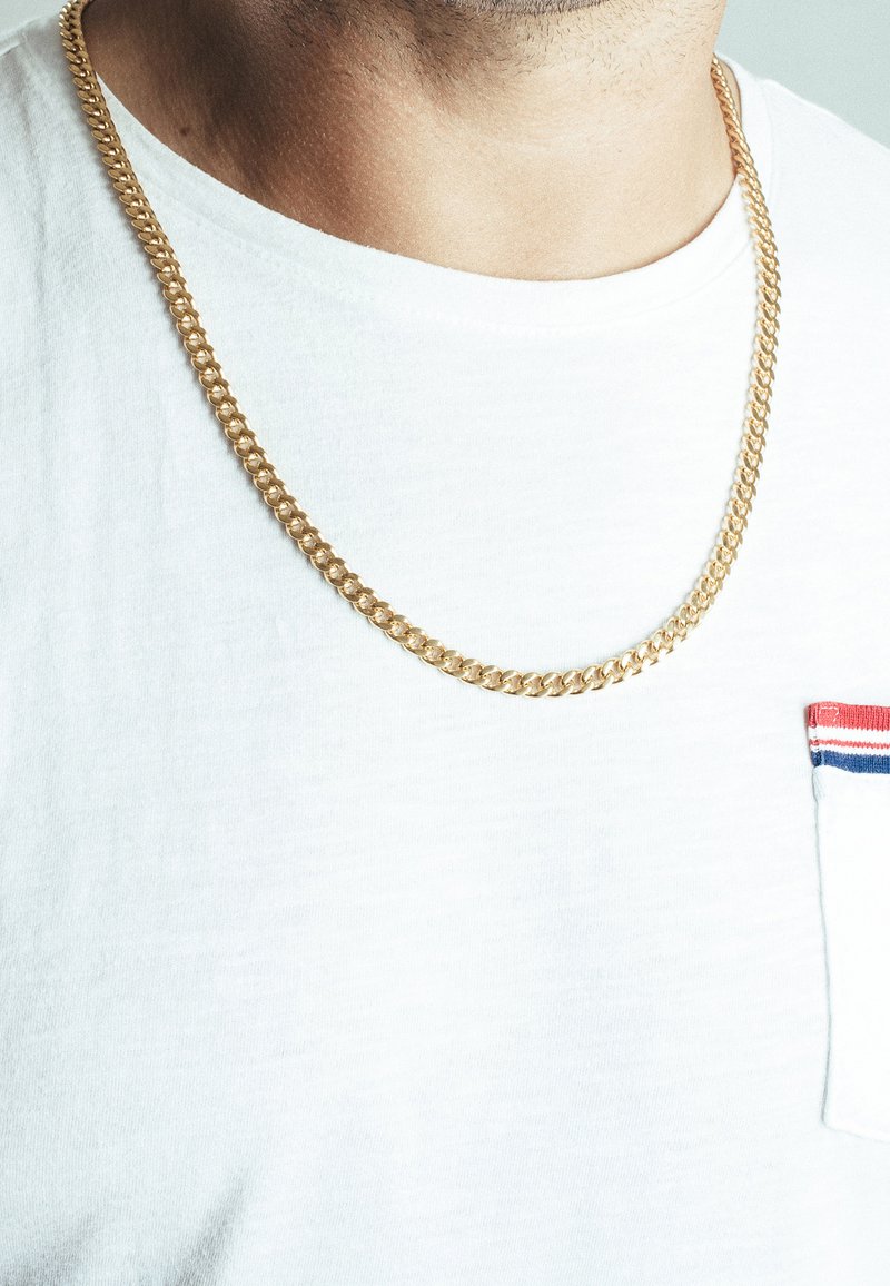 Gouden ketting met in elkaar grijpende schakels, rustend over een wit T-shirt met een klein zakje met rode en blauwe accenten.