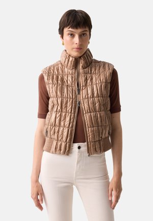 Kvinde iført en beige quiltet ærmeløs vest over en brun top med korte ærmer og hvide bukser med høj talje, vendt mod fronten.