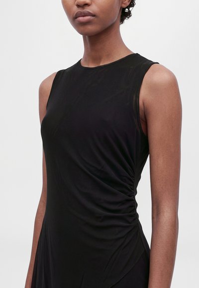 Modèle portant une robe noire sans manches avec du tissu froncé à la taille, sur un fond clair uni.