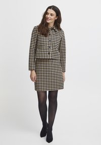 Pruun ja must houndstooth mustriga kahest osast koosnev komplekt, mis sisaldab kohandatud lühikest jakki ja liibuvat mini seelikut, millele on lisatud mustad sukkpüksid ja kontsad.