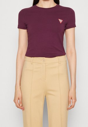 Kobieta ubrana w dopasowany fioletowy t-shirt z krótkim rękawem z małym czerwonym logo Guess na piersi oraz beżowe spodnie z wysokim stanem.