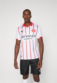 Camisola de futebol branca com riscas verticais vermelhas, gola vermelha e texto preto "ETIHAD AIRWAYS." Usada com calções pretos, exibindo um design desportivo.