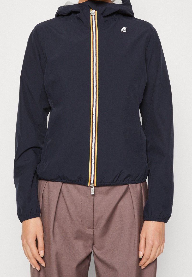 Veste à capuche bleu marine avec fermeture éclair frontale affichant des rayures jaunes, blanches et rouges, portée avec un pantalon marron taille haute.