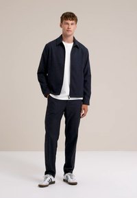 Marinblå zip-up jacka över en vit t-shirt, matchad med marinblå byxor och vita sneakers med svarta detaljer. Enkelt, stilrent design.