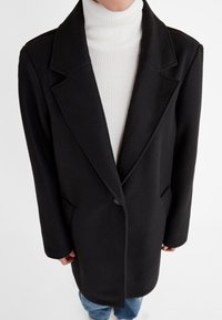 Persoon in een zwart op maat gemaakt coat over een witte geribde turtleneck trui en blauwe jeans, staand tegen een witte achtergrond.