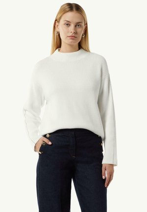 Weißer gerippter Strickpullover mit hohem Kragen und langen Ärmeln, kombiniert mit dunklen Jeans, die goldfarbene Hardware-Details aufweisen.