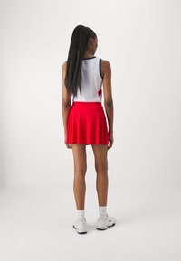 Röd sportkjol med veck, tillsammans med en vit linne med blå och röda detaljer. Modellen bär vita sneakers med logotyp och strumpor.
