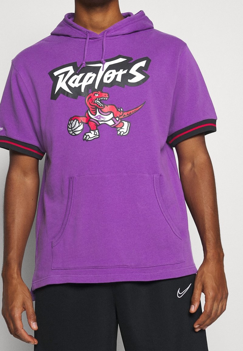 Sweat à capuche violet à manches courtes avec le logo des Raptors montrant un dinosaure rouge en train de dribbler un ballon de basket, porté avec un pantalon noir Nike.