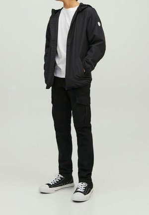 Giacca nera con cappuccio e chiusura lampo, t-shirt bianca sotto, pantaloni cargo neri e sneakers nere con lacci e dettagli bianchi.