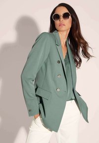 Cyaan blazer met dubbele rij knopen, zilveren knopen en zijzakken, gelaagd over een kanten top, gecombineerd met witte broeken.