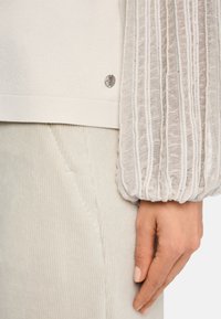Helles graues Sweatshirt mit strukturierten, plissierten Ärmeln. Verfügt über einen glatten Körper und ein silbernes Metall-Logo-Detail am Saum. Kombiniert mit hellen Cordhosen.