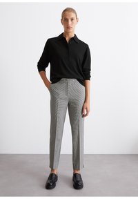 Polo shirt noir avec un col et des boutons, associé à un pantalon à carreaux gris avec des fentes latérales. Les chaussures sont des mocassins noirs.