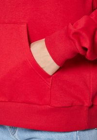 Czerwona bluza z kangurzą kieszenią, z prążkowanymi mankietami i dołem. Wykonana z miękkiego materiału, zawiera widoczne szwy i gładką fakturę.