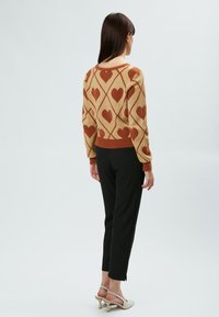 Beige Strickpullover mit braunem Herzmuster und Rautenmuster; gerippte Bündchen und Saum. Kombiniert mit schwarzen eng anliegenden Hosen und beigen High Heels.