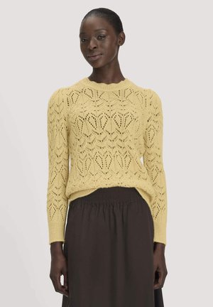 Femme portant un pull en maille jaune à motifs et une jupe sombre taille haute, debout devant un fond clair uni.