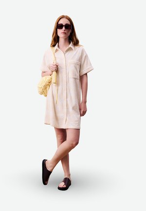 Femme aux cheveux roux portant une robe chemise crème à fleurs, des lunettes de soleil foncées, des sandales noires à enfiler et un sac à épaule jaune froncé.
