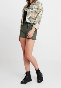 Lyhythrintainen takki, jossa on kamouflagemotiivi, musta graafinen t-paita, vihreät teksturoidut shortsit nyörillä ja mustat nauhalliset saappaat. Yksinkertainen malli, rento ilme.