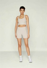 Ung kvinne med kort hår står iført en beige ribbet crop top og matchende høytlivsshorts med hvite sokker og sneakers.