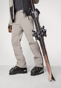 Person wearing beige ski pants and black ski boots holding a pair of black and red skis upright on a gray floor.