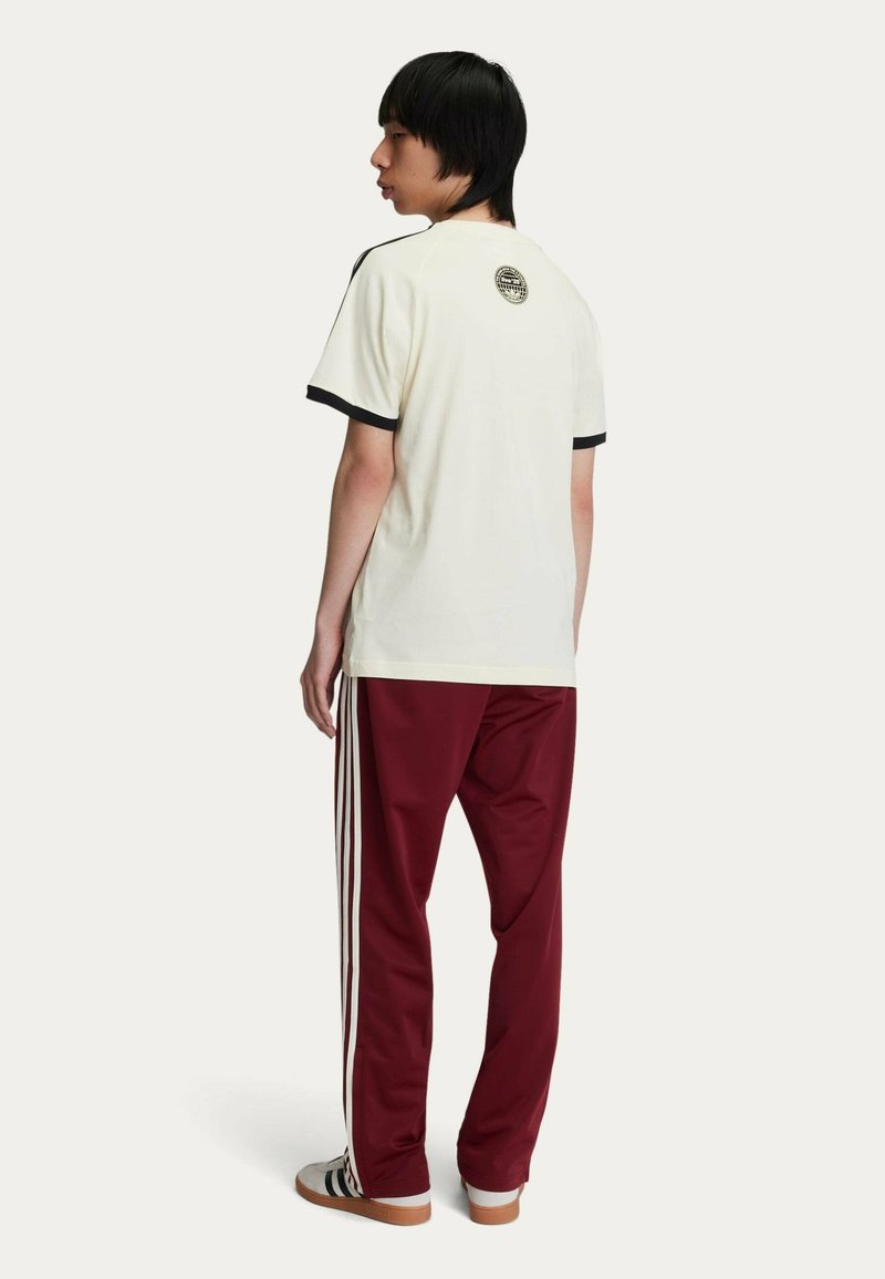 T-shirt crema a maniche corte con bordi neri, abbinato a pantaloni bordeaux con strisce bianche sui lati. Collo aperto, vestibilità casual.