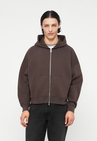 Sweat à capuche zippé marron avec une grande capuche, des poches avant et un design court. Fabriqué en tissu doux avec une texture lisse, avec des poignets côtelés.