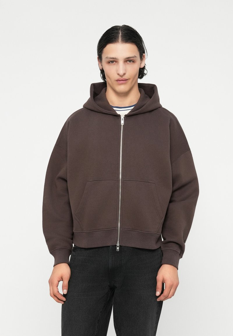 Sweat à capuche zippé marron avec une grande capuche, des poches avant et un design court. Fabriqué en tissu doux avec une texture lisse, avec des poignets côtelés.