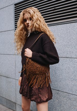Une femme aux longs cheveux blonds et bouclés se tient près d’un mur en pierre. Elle porte une tenue sombre et un sac à bandoulière à franges, imprimé léopard.