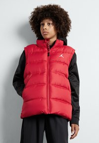 Gilet rembourré rouge avec une fermeture éclair à l'avant, présentant un design matelassé et un logo sur la poitrine gauche, porté par-dessus un haut à manches longues noir.