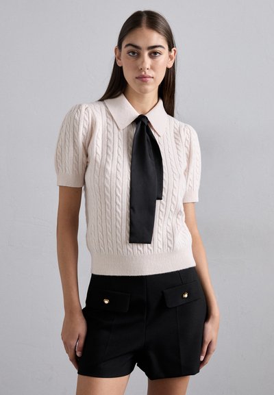 Pulover tricotat în bej deschis cu model împletit, mâneci scurte cu volum și o fundiță din satin negru. Asortat cu pantaloni scurți negri cu talie înaltă, prevăzuți cu buzunare.